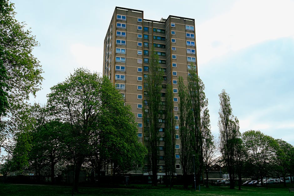 An upward-facing view of a tall, multi-storey residential building constructed with brown brickwork, featuring numerous small, rectangular windows and several small balconies with metal railings on th